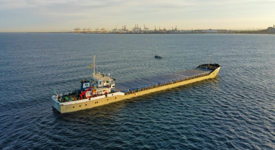 Aerial view of a cargo ship sailing near a busy industrial port at sunset.